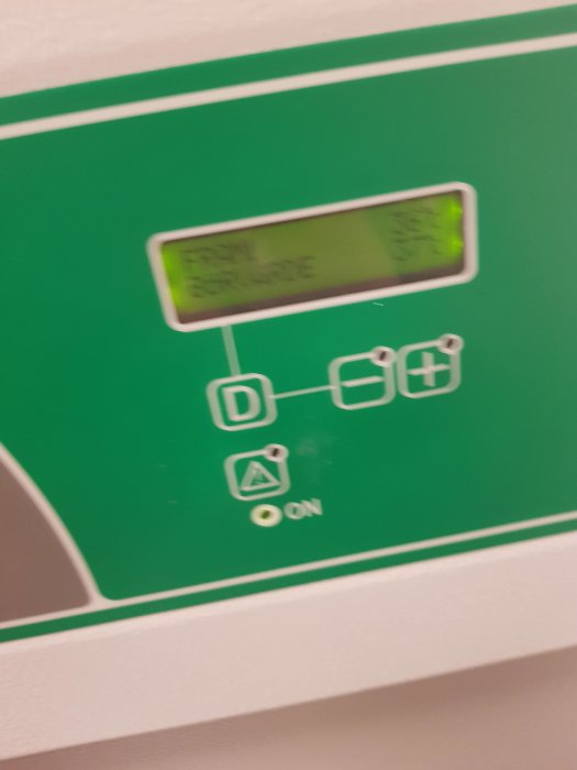 Grönt elektroniskt kontrollpanel med suddig LCD-display och symboler för strömställare och batterier.