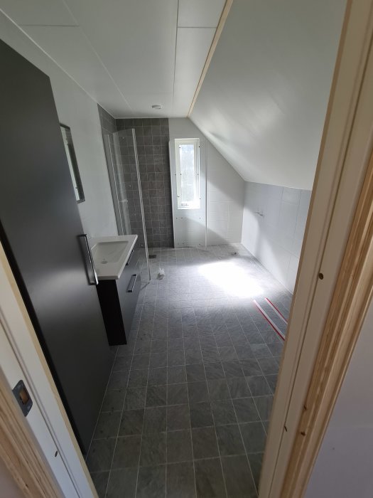 Nybyggt badrum med sluttande tak, grå kakelväggar och golv, vit handfat och ett fönster.