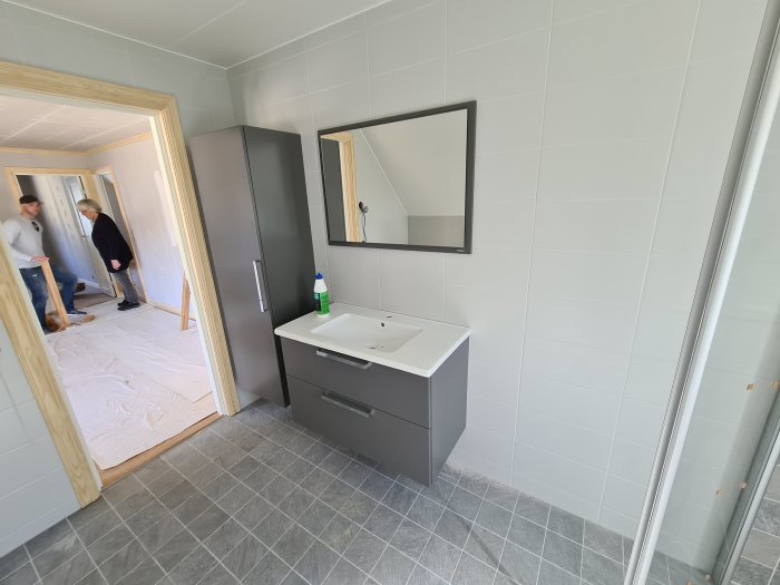 Ett pågående badrumsprojekt med grå badrumsskåp och spegel, vita kakelväggar och grått klinkergolv, två personer i bakgrunden.