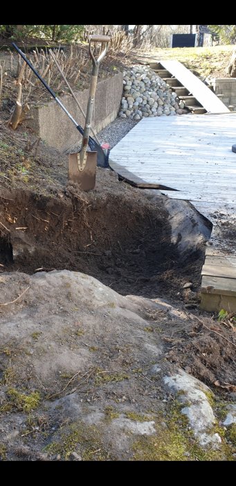 En trädgårdsspade står lutad mot en stor sten intill en nedgrävd del av en altan, indikerar byggprojekt med stenspräckning.