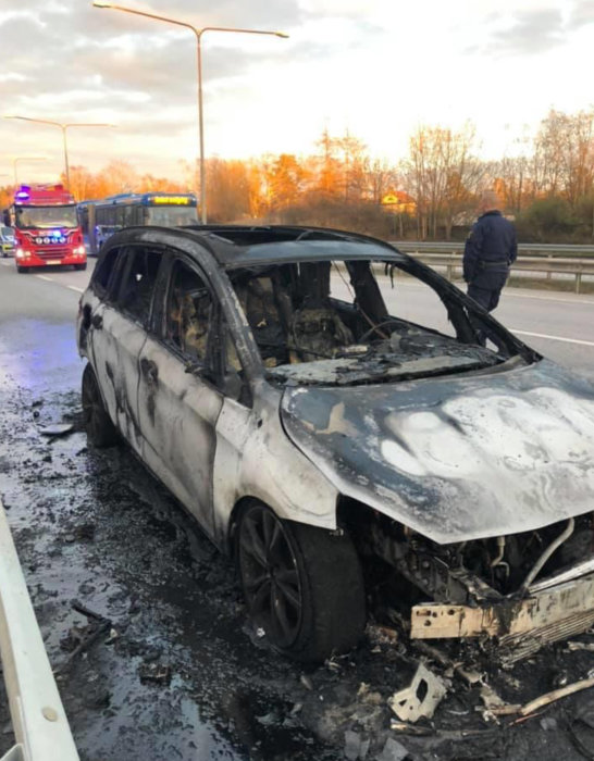 Utförbränd bil vid vägkanten med brandbil i bakgrunden och en person som observerar skadorna.