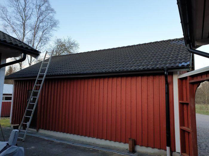 Utsidan av en carport ombyggd till hemmagym med röda träväggar och svart tak av tegelpannor, med en stege lutad mot väggen.