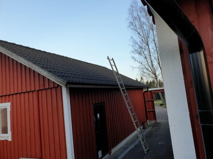 Rödmålad carport med anslutande förråd och stege mot taket, tegelpannor synliga, i ett hemmiljö.