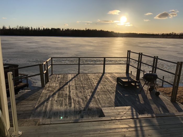 Altan med utsikt över en frusen sjö vid solnedgång, omgiven av ett enkelt räcke, med grill och sittmöbler.