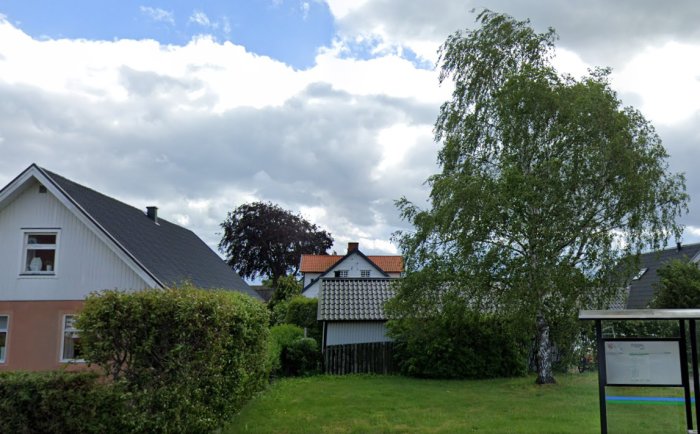 Hus med vita fasader och solcellspaneler på taket, beläget sydväst om ett stort grönt träd, under en molnig himmel.