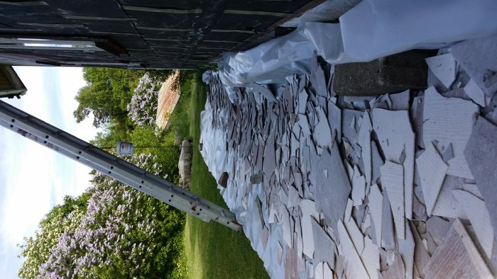 Sönderbrutna eternitskivor ligger på marken bredvid en stege, med tjärpapp synlig under skivorna och grönska i bakgrunden.