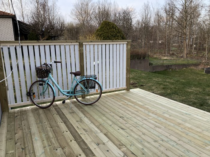 Nybyggt trädäck med ett enkelt vitmålat staket och en turkos cykel, gräsmatta i bakgrunden.