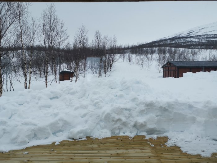 Vinterlandskap med tjockt snötäcke framför terrass, små stugor och fjäll i bakgrunden.