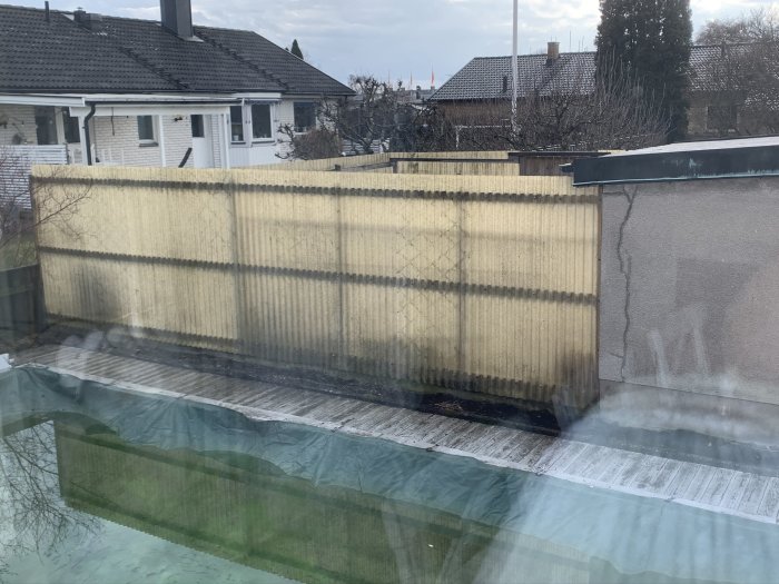 Gammal plastvägg längs med en altan som vetter mot en grå husvägg och trädgård.