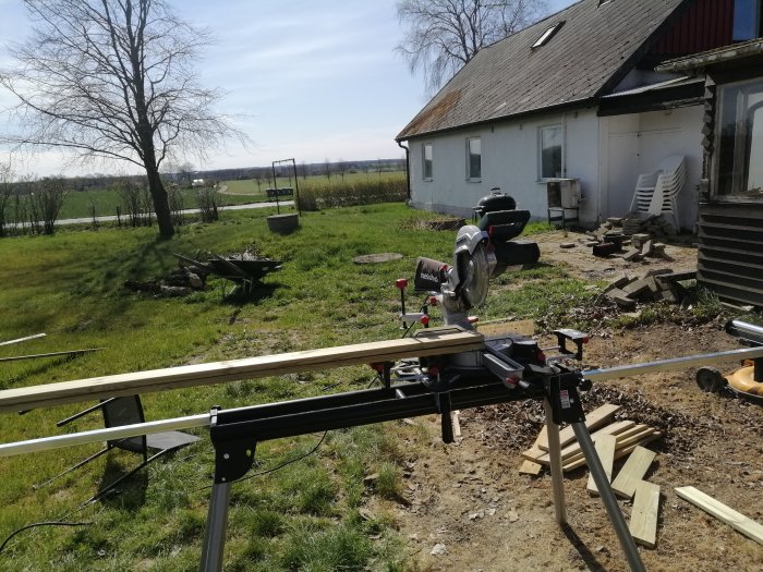 En arbetsplats utomhus med såg och träplank vid ett under construction vedförråd nära ett hus.