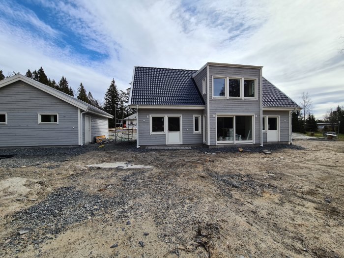 Nyligen byggt hus med grå fasad och svart tak intill en garagebyggnad under ett molnigt himmel.