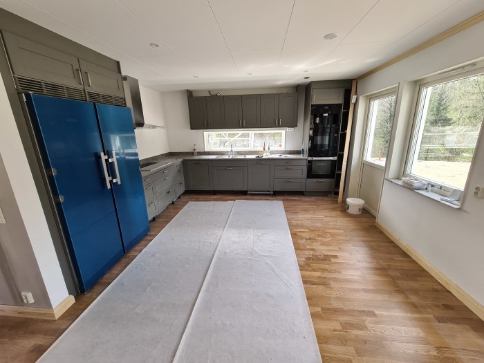 Modernt kök med grå skåp, blå kylskåp och parkettgolv, delvis täckt av skyddsplast.