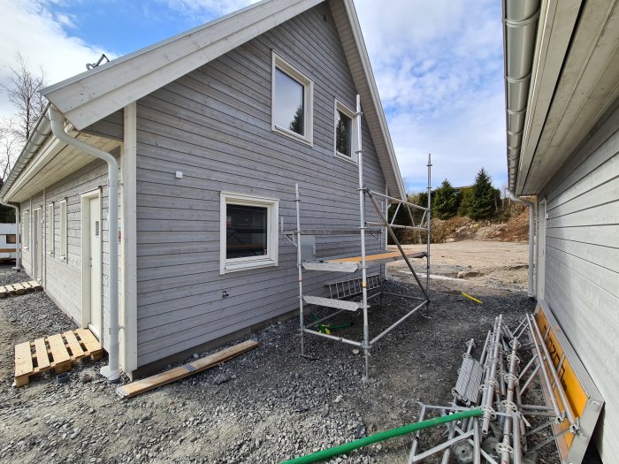Nybyggd grå träfasad med vita fönster och dörrar, byggställning vid sidan, och byggmaterial på marken.