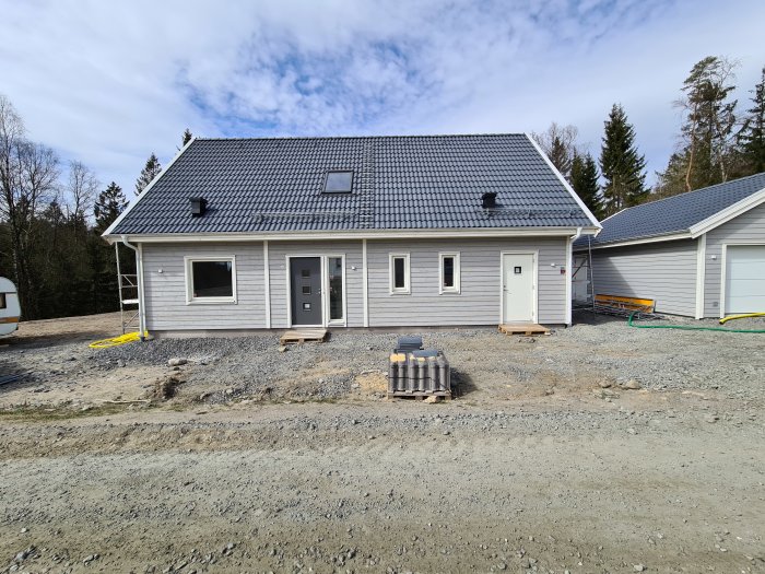 Nybyggt enplanshus med anslutet garage under en klarblå himmel, omgivet av grus och byggmaterial.