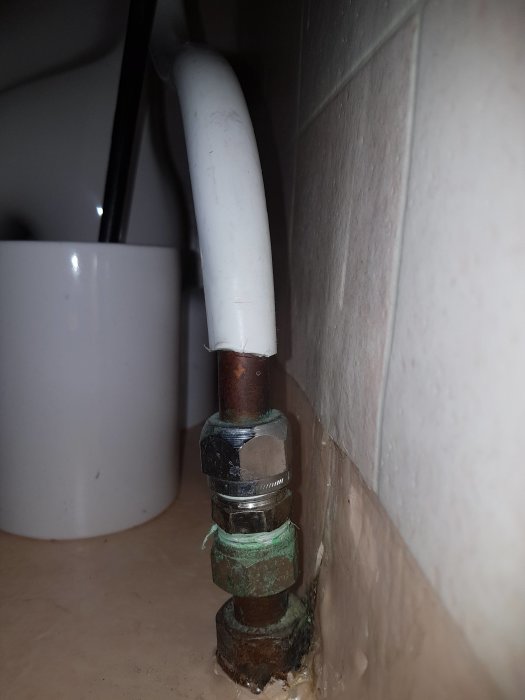 Kopparfärgad mutter anliggande vägg, med ett mått på 12 mm upp till en blank mutter, bakom vit rörledning.