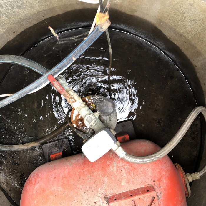 Bild på en öppnad brunn med röd pump och rörledningar inne i brunnen, vatten syns i bakgrunden.