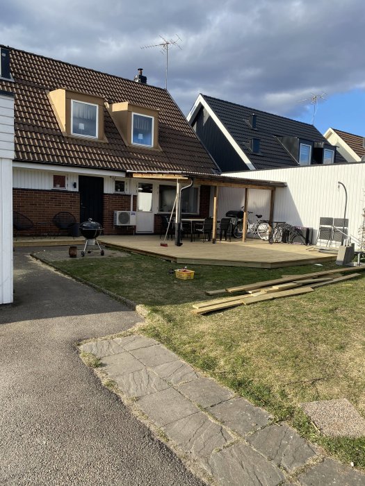 Bygge av trätrall framför hus, plankor på gräset, trädgårdsmöbler och grill på verandan.