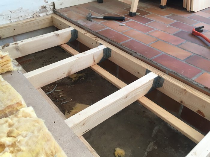 Gulvisolering og træreglar fjernet i en kælder, blotlagt under lerfliser; værktøj synligt på fliseoverfladen.
