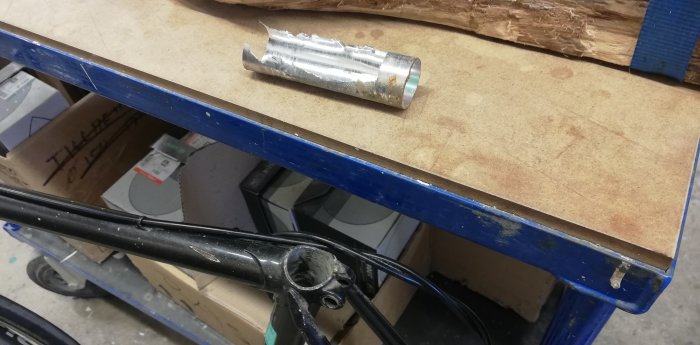 Utburrad aluminiumsadelstolpe på arbetsbänk bredvid cykelram med fast roterande sadelstolpsklämma.