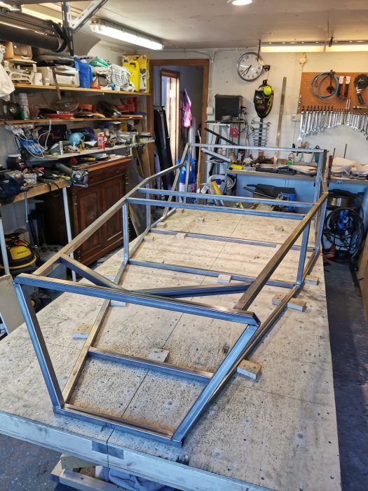 Metallramar på arbetsbord i ett rörigt garageverkstad med verktyg och cyklar i bakgrunden.