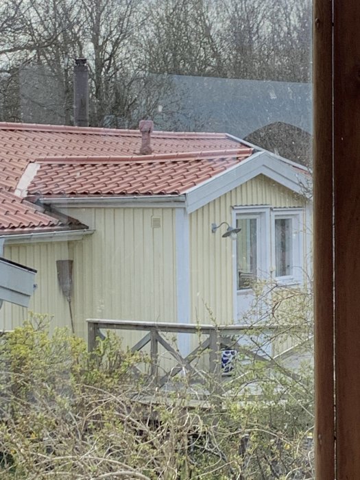 Matt rött plåttak på gult hus sedd genom ett fönster, med träd och buskar i förgrunden.