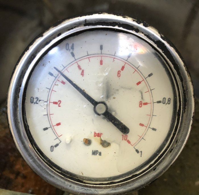 Smutsig manometer med visare vid 0,5 bar, skalor märkta i både bar och MPa, ovanpå en skrovlig yta.