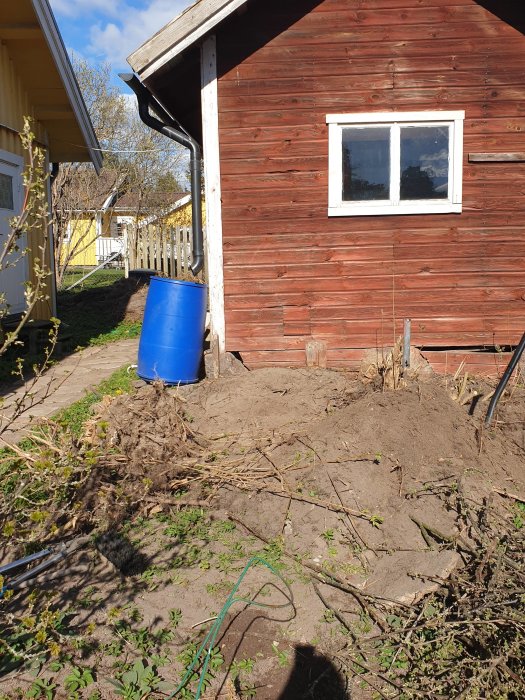 Ommålning av stuga med blå tunna och spade vid grävd jord i en solig trädgård.