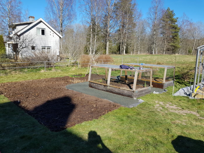 Trädgård med ett område grävt för bergkross, intill odlingslådor och med ett hus i bakgrunden.