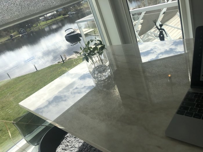 Arbetsplats med dator på bord och utsikt över stilla vatten med båt och spegelblank yta.