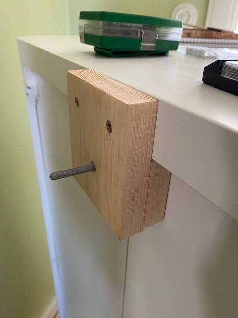 Träkloss fästad på baksida av vit hyllskiva med en spikplugg som sticker ut.