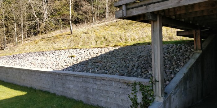 Stödmur av gråa betongblock med grus och stenar framför och ett skogsområde i bakgrunden vid sidan av ett hus.