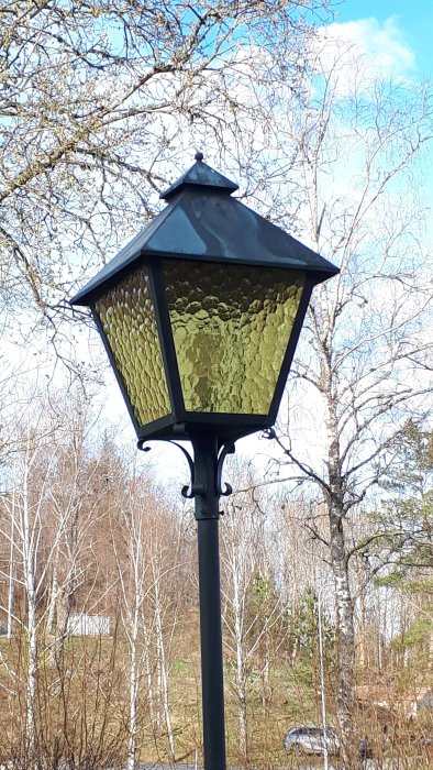 Lyktstolpe utomhus med intakt gulaktigt glas och naturbakgrund.