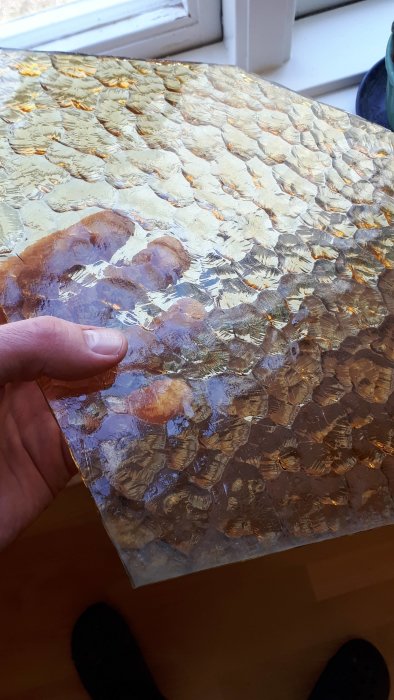 Hand håller ett brutet, strukturerat glas med mönster, troligen från en lyktstolpe, nära ett fönster.