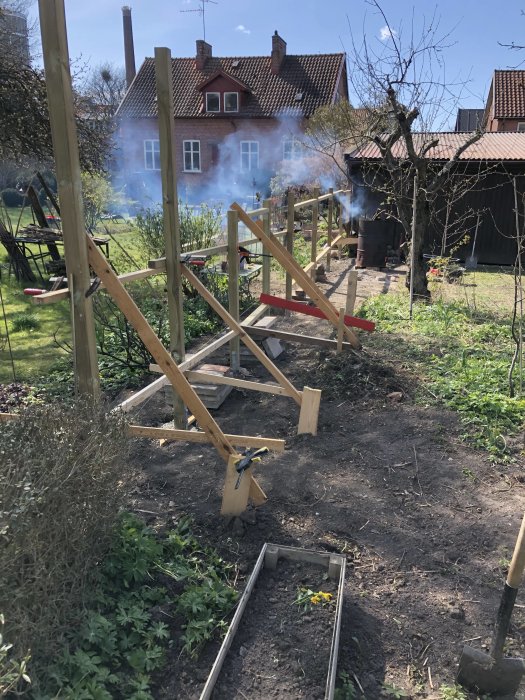 Nyuppsatta trästolpar i trädgård med hus och rökmoln i bakgrunden.