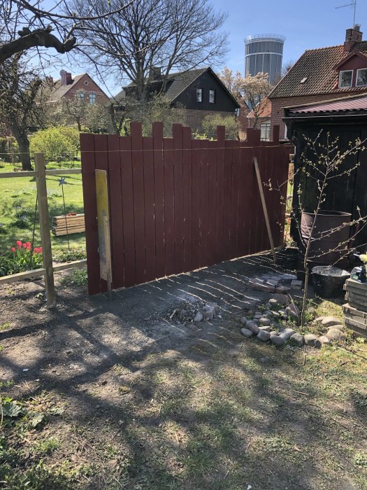 Delvis uppfört trästaket med nya rödmålade plank och stolpar i en trädgård, med grannhus och träd i bakgrunden.