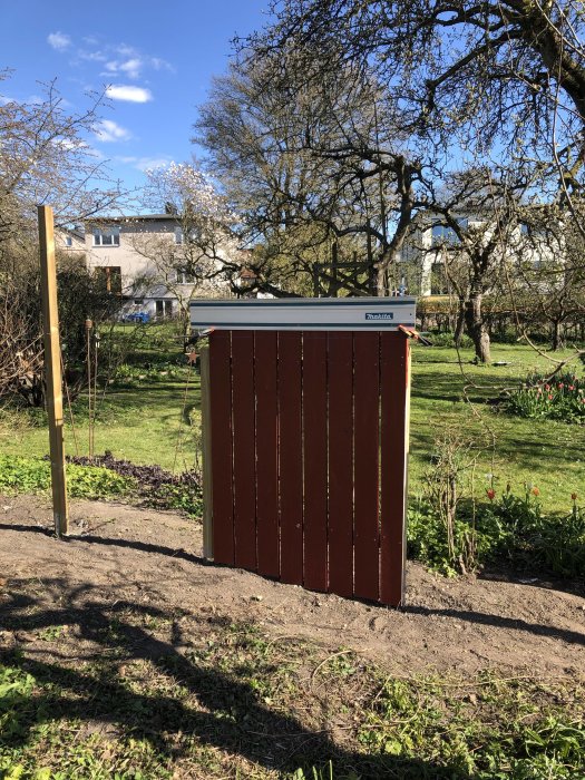 Stolpar och nyuppsatta röda träplank på plats i ett trädgårdsstaket, verktygslåda och grönska i bakgrunden.