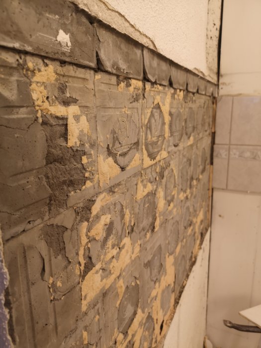 Væg med fjernet flisebelægning, hvor rester af klæbemiddel og mørtel er synlige, hvilket skaber en ujævn og ru overflade.