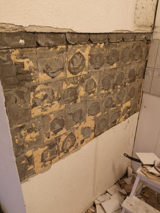 Væg med fjernet fliser, der afslører skrovlig overflade og rester af mørtel. Fliser og værktøj ligger på gulvet.