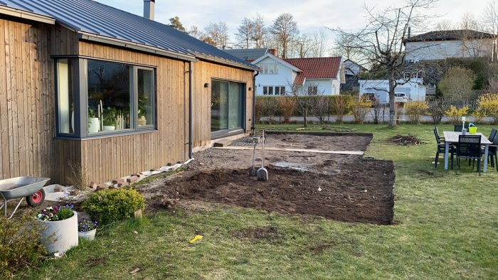 Uppmärkt trädgård för altanbygge bredvid trähus, skottkärra och trädgårdsredskap syns.