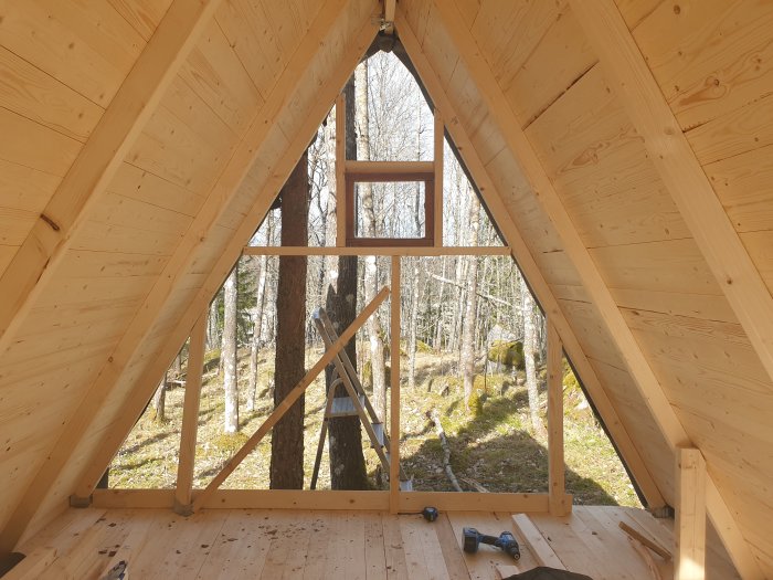 Interiör av en stuga under konstruktion med triangulärt fönster och synligt timmerarbete, omgiven av skog.