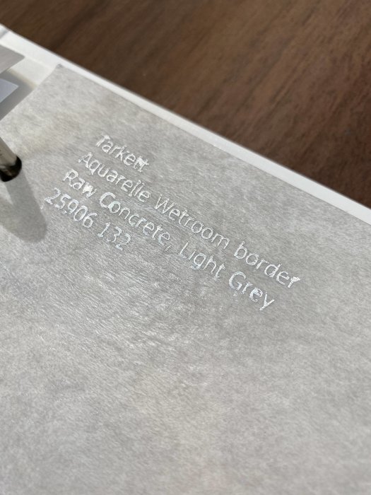 Etikett med texten "Tarkett Aquarelle Wetroom border Raw Concrete Light Grey" mot en bakgrund av mönstrad matta.