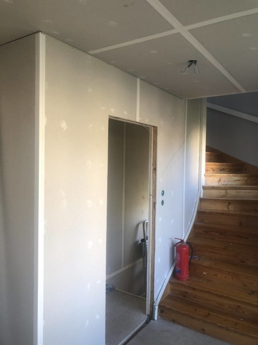 Rum under renovering med spacklade väggar nära trätrappa och öppen dörr, första spackelvarvet med synliga skarvremsor.