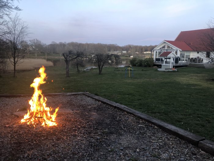 Eld i en trädgård vid skymning med ett hus och sjö i bakgrunden.