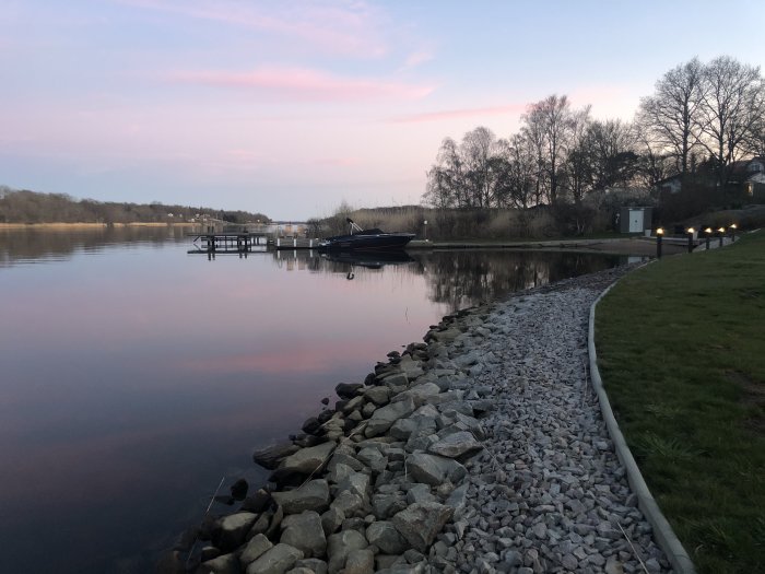 Skymningsvy över en stilla sjö med rosa himmel, stenstrand och en båtbrygga med båt.