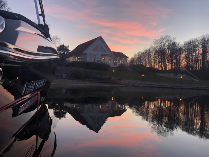 Kvällshimmel i nyanser av rosa och blå över en stilla sjö med spegling av ett hus och träd, framkant av en båt synlig.