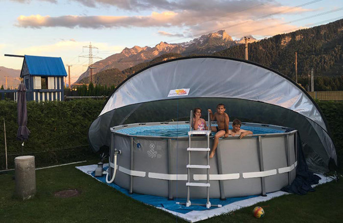 Halvrund pooltält över en utomhuspool med barn som sitter vid kanten, berg i bakgrunden.