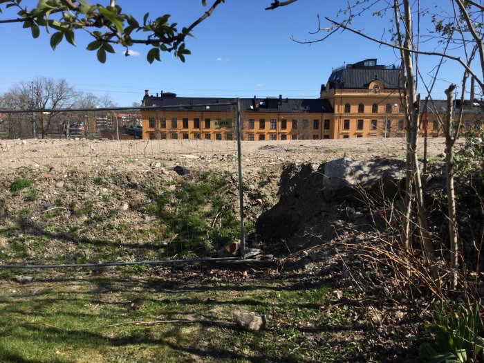 Ojämn tomtyta med berg och utsikt mot byggnad, nedsågade träd, oordnad gräns mot grannfastighet.