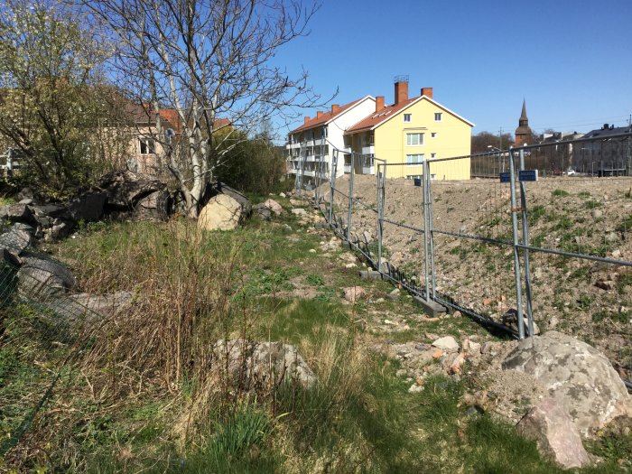 Ojämn tomt med berggrund, avsågade träd, byggstängsel och bakgrund av hus och himmel.