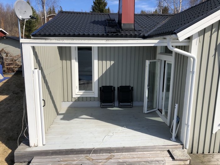 Utsikt över en husveranda med vitmålade väggar, två svarta stolar, öppna terrassdörrar och ett tak med röd skorsten.