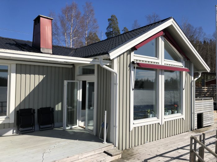 Modernt grått hus med stora fönster och en terrass med utsikt över en sjö, under en klarblå himmel.